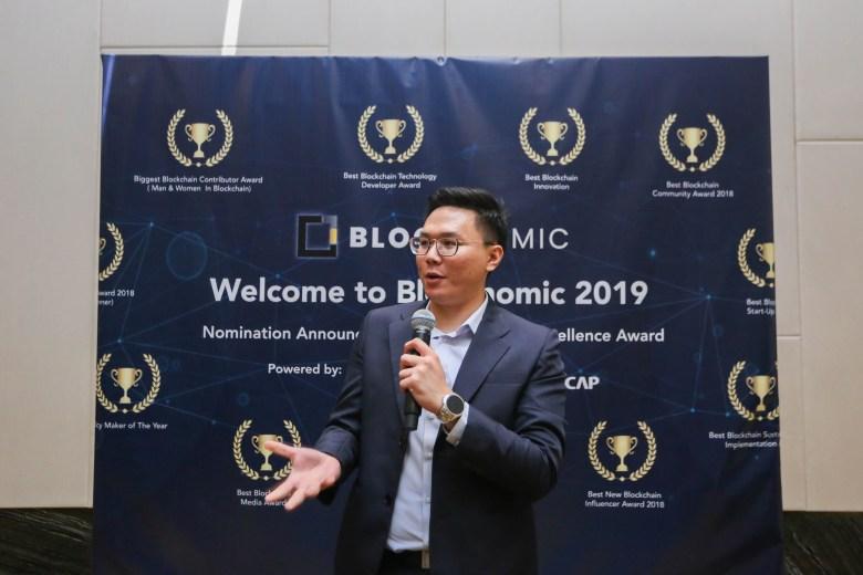 Trivechain nominated for bloconomic excellence award at the summer bloconomic sumit 2019 Man speaking with a bloconomic image in the background